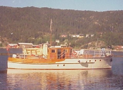 En båt (Hauka) med styrhus i tre og hvitt skrog ligger på stille vann en solskinnsdag. Båten speiles i vannet. I bakgrunnen skogkledte fjellsider med noe bebyggelse. 