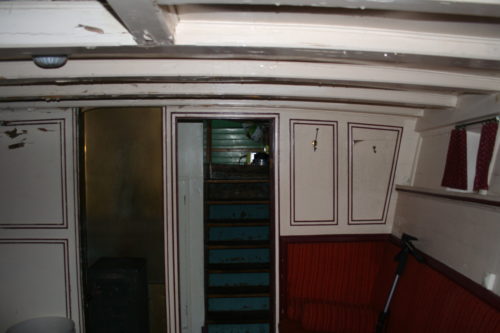Salong hvor veggene er grå med rustrødt brystningspanel. Til venstre i bildet er en teakdør, ved siden av en døråpning hvor vi ser en trapp opp.